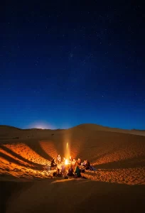 People gathered around a campfire under an indigo sky in the Sahara desert against sand dunes