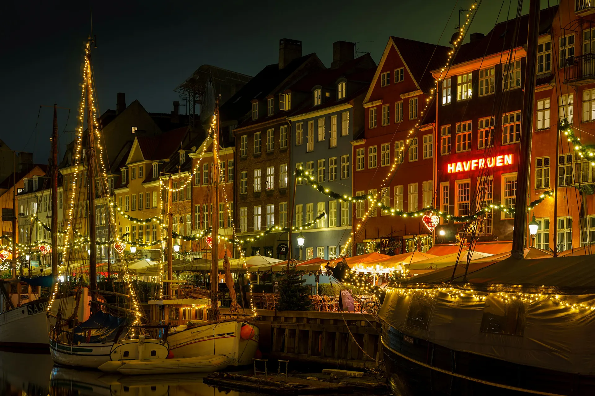 Boats moored in front of traditional colorful Danish row houses. The boats and houses are decorated with Christmas lights. The photo is taken at night and the setting looks cozy and cheerful.