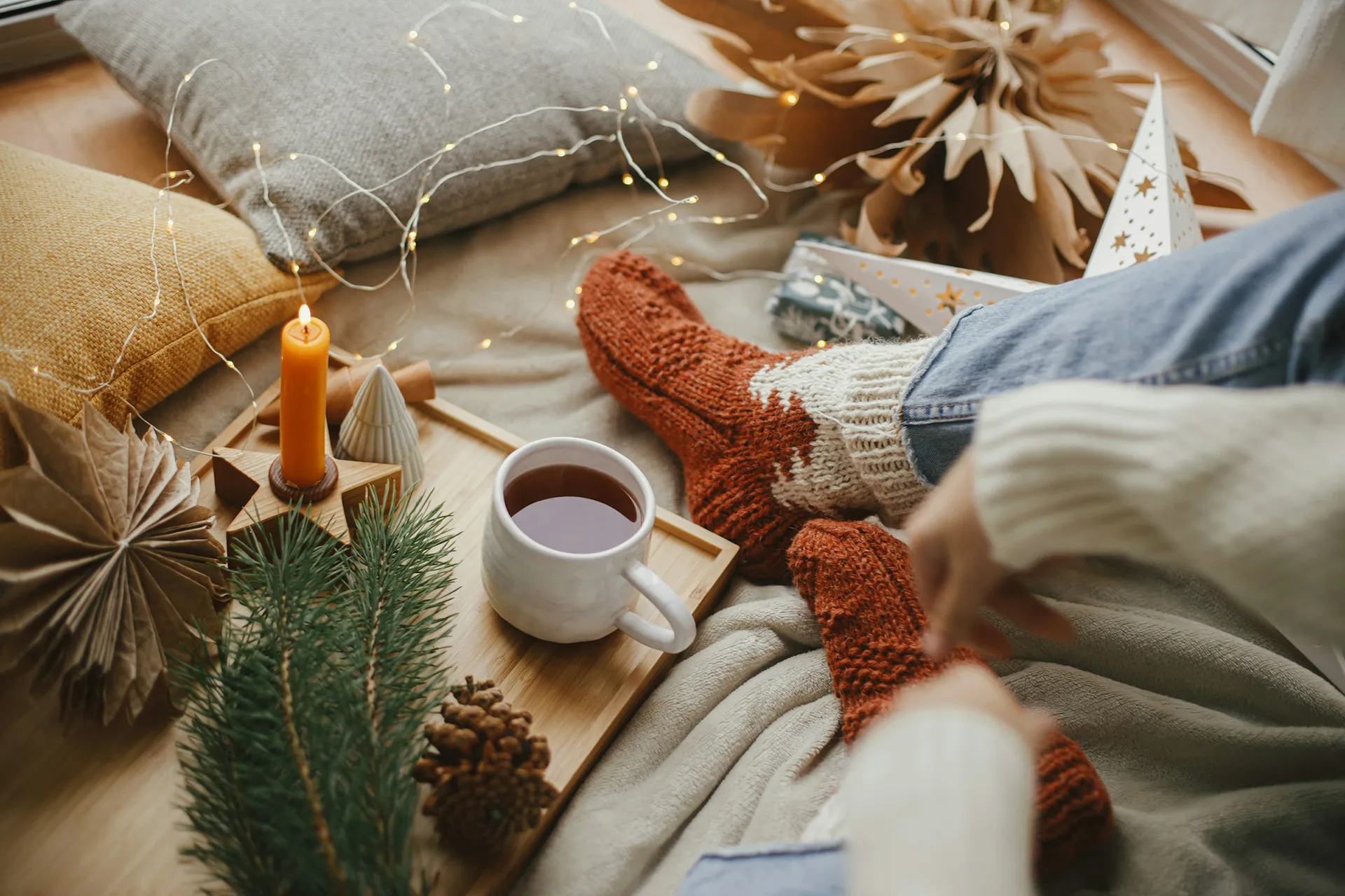 Overhead shot of person's legs sitting on the floor, wearing cozy socks and a sweater. The person has a mug of tea next to them and is surrounded by cozy looking decor.