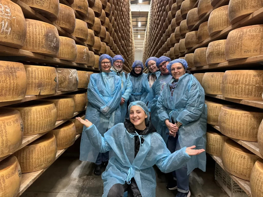 Group of tourists in blue protective gear posing in between rows of Parmigiano Reggiano cheese in Emilia Romagna